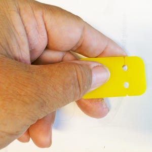 May include: A yellow plastic squeegee with two holes. The squeegee is being held by a hand.