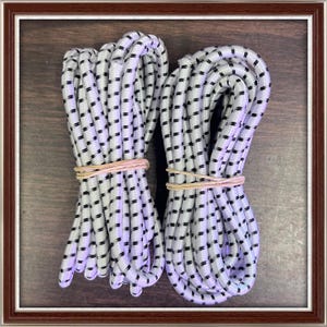 May include: Two bundles of white and black patterned bungee cords, secured with tan rubber bands. The cords are tightly coiled and presented against a dark wood-grain background within a brown framed border.