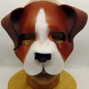 May include: A brown and white leather dog mask with black nose and white fur details. The mask is on a wooden mannequin head.