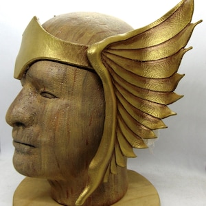 May include: A gold-colored leather headband with a winged design. The headband is worn on a mannequin head.