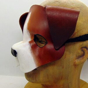 May include: A brown and white leather dog mask with a black elastic strap. The mask has a white nose and brown ears.