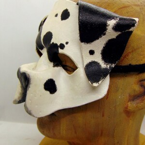 May include: A white and black dalmatian dog mask with black spots. The mask has a cutout for the eyes and a nose.