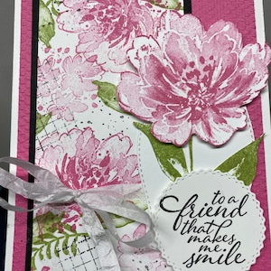 May include: A white card with a pink and green floral pattern. A white circle with a black border and the text "to a friend that makes me smile" is on the right side of the card. A white ribbon is tied around the card.