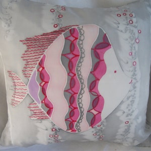 Puede incluir: Una almohada blanca con un dise&ntilde;o de pez rosa y gris. El pez tiene un cuerpo estampado en rosa, morado y gris y una cara blanca con una boca rosa.