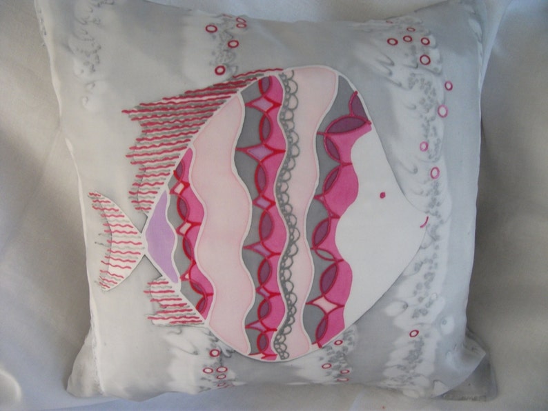 Puede incluir: Una almohada blanca con un dise&ntilde;o de pez rosa y gris. El pez tiene un cuerpo estampado en rosa, morado y gris y una cara blanca con una boca rosa.