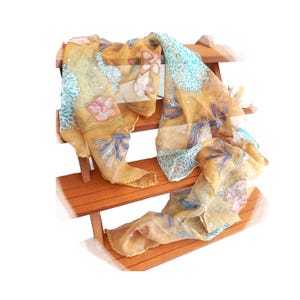 May include: A sheer, mustard-yellow scarf with a floral pattern of pink, blue, and white flowers and accents. The scarf is draped over a wooden display stand, showcasing its flowing fabric and design.