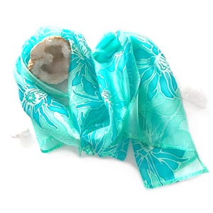 May include: A turquoise silk scarf with a floral pattern in shades of blue and white. The scarf is draped over a geode and a crystal cluster. The scarf's design features large, stylized flowers.