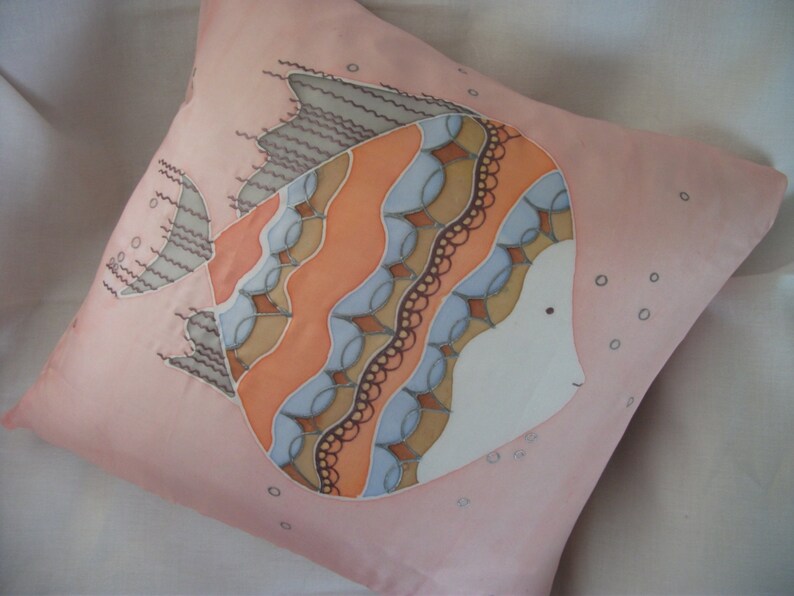 Puede incluir: Una almohada rosa con un dise&ntilde;o pintado a mano de un pez estilizado con detalles en naranja, marr&oacute;n y azul. El pez est&aacute; rodeado de peque&ntilde;os c&iacute;rculos.