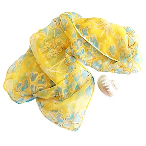 Puede incluir: Un pañuelo amarillo con un patrón repetitivo de corazones azul claro delineados en oro, con el texto "Sending you a box of kisses" impreso en la tela. Junto al pañuelo hay un pequeño objeto metálico plateado con forma de concha estriada.