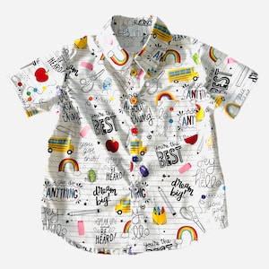 Può includere: Camicia bianca a maniche corte con bottoni con una stampa colorata di scarabocchi a tema scolastico su tutta la superficie, tra cui arcobaleni, autobus scolastici, matite e le parole "You're the Best", "Dream Big" e "Speak Up and Be Heard!"