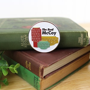 May include: A white enamel pin with a red, yellow, and green design of three ceramic planters. The text "The Real McCoy" is printed in black on the pin.