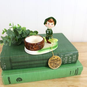 May include: A ceramic figurine of a pixie wearing a green hat and outfit, positioned next to a brown basket-weave candle holder on a green base. A gold-colored metal tag hangs from the figurine, inscribed with the text: Helpful Kitchen Pixie Says, Light Candle To Freshen Air.