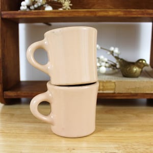 May include: Two stacked, light peach-colored ceramic mugs with rounded handles. The mugs are on a wooden surface, with a wooden shelf in the background. A small brass bird figurine and decorative white berries are also visible.
