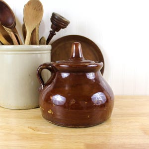 May include: A brown glazed ceramic jar with a lid and a handle. The jar is sitting on a wooden surface.