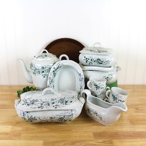 May include: A collection of white ceramic dinnerware featuring a dark green botanical floral pattern. The set includes a lidded teapot, a lidded rectangular tureen, a gravy boat, several small cups, and an oval serving platter arranged together.