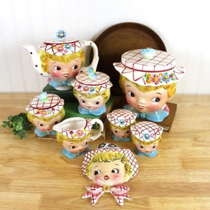 May include: A ceramic tea set featuring stylized faces with blonde hair and blue bows. The set includes a teapot, a large lidded jar, a creamer, three smaller lidded jars, and a wall plaque, all decorated with red grid patterns and floral accents.