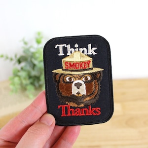 May include: Embroidered patch featuring Smokey Bear wearing a ranger hat. The patch has the words "Think" and "Thanks" in white and red, respectively. The word "Smokey" is on the hat. The patch is on a black background.