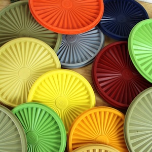 May include: A collection of round, plastic container lids in various colors including orange, dark blue, olive green, yellow, bright green, red, and grey. Each lid features a central circular design with radiating sunburst-like ridges.