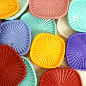 May include: A collection of square and rounded plastic storage container lids in various colors including red, yellow, purple, blue, teal, pink, and white. Each lid features a central circular indentation and a radiating sunburst pattern.