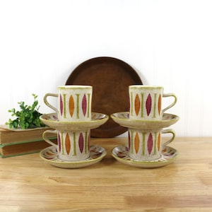 May include: Set of four ceramic coffee cups and saucers. The cups and saucers have a speckled beige background with vertical orange, green, and maroon designs. A large, round, wooden tray is in the background.
