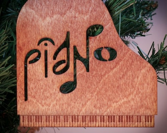 Op de afbeelding: Houten kerstversiering in de vorm van een piano met het woord "piano" en uitgesneden muzieknoten. De versiering heeft een rij toetsen aan de onderkant.