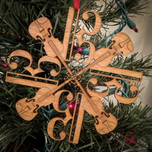 Könnte beinhalten: Weihnachtsbaumschmuck aus Holz in Form einer Schneeflocke mit Noten und Violinschlüsseln. Der Schmuck ist in Brauntönen bemalt und zeigt ein Design mit vier Violinen.