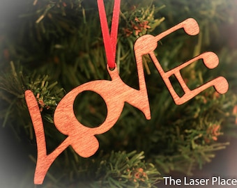 Op de afbeelding: Een houten kerstversiering in de vorm van het woord "LOVE" met muzieknoten. De versiering is in een roodbruine kleur gebeitst en heeft een rood lint.