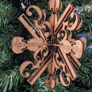 Puede incluir: Adorno navideño de madera con forma de copo de nieve que presenta un diseño de violines y notas musicales. El adorno es de madera marrón claro y tiene una cinta verde para colgarlo. El diseño es intrincado y detallado.