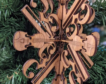 Puede incluir: Adorno navideño de madera con forma de copo de nieve que presenta un diseño de violines y notas musicales. El adorno es de madera marrón claro y tiene una cinta verde para colgarlo. El diseño es intrincado y detallado.