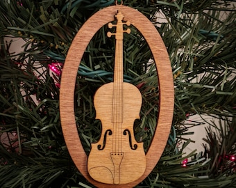 May include: A wooden Christmas ornament shaped like a violin. The ornament is brown and has a cutout design of a violin. The ornament is hanging from a green ribbon on a Christmas tree.