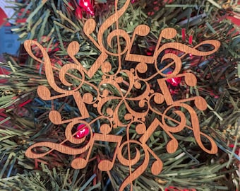 Pode incluir: Um enfeite de Natal de madeira em forma de floco de neve com notas musicais recortadas. O enfeite é marrom e está pendurado em uma árvore de Natal verde.