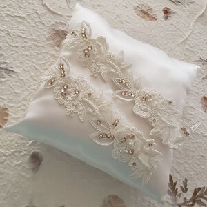 May include: Two white lace garters with delicate floral designs and sparkling rhinestones, displayed on a white satin pillow.