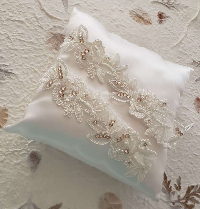 May include: Two white lace garters with delicate floral designs and sparkling rhinestones, displayed on a white satin pillow.