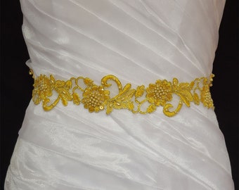 May include: A gold-colored, embroidered floral belt with pearl accents. The belt is made of a delicate, open-weave fabric and features a repeating pattern of flowers and leaves. The belt is shown against a white, pleated fabric.
