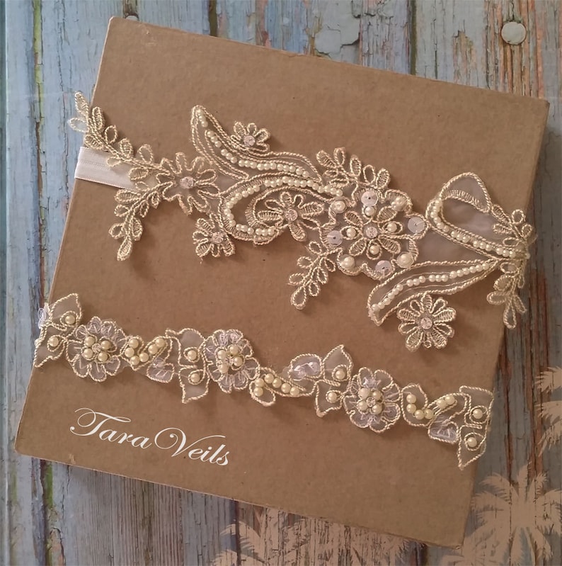 May include: Ivory lace bridal garter set with floral and pearl embellishments. The set includes a larger piece and a smaller garter, both with delicate detailing. The garter set is presented on a brown box with the text "Tara Veils".