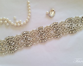 May include: A white satin ribbon with a crystal and pearl embellishment. The embellishment features a floral design with a silver finish. The ribbon is tied in a bow and the text "Tara Veils" is visible on the ribbon.