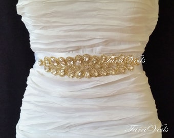 May include: A white satin bridal sash with a gold rhinestone embellishment. The embellishment features a cluster of clear rhinestones in a leaf-like design. The sash is shown on a white dress.