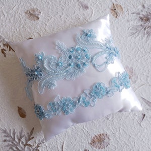 May include: A white satin pillow with delicate blue lace trim and pearl accents. The lace features a floral design with a vintage aesthetic.