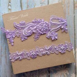 May include: Two lavender lace garters with pearl embellishments. The top garter features a floral design with a larger flower in the center. The bottom garter has a more delicate floral design with pearls throughout.
