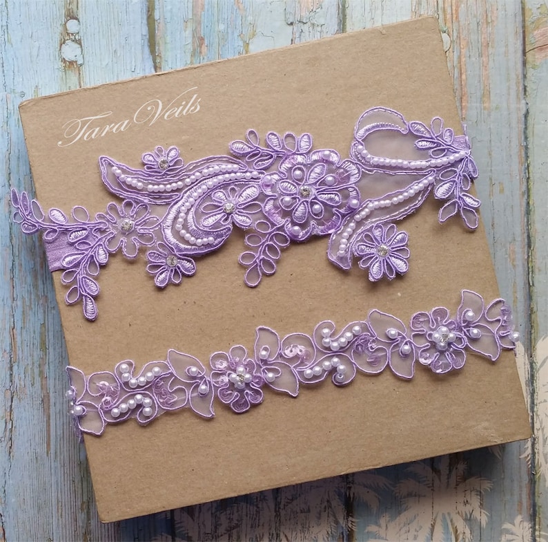 May include: Two lavender lace garters with pearl embellishments. The top garter features a floral design with a larger flower in the center. The bottom garter has a more delicate floral design with pearls throughout.