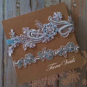 May include: Two delicate lace garters with light blue and silver embellishments. The garters are adorned with pearls and sequins. The text "Tara Veils" is visible in the bottom right corner.
