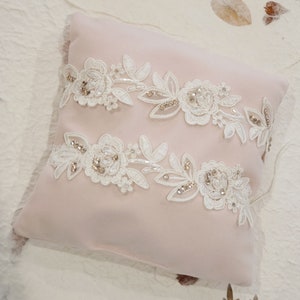 May include: A blush pink pillow with two white lace appliques featuring floral designs and sparkling embellishments.