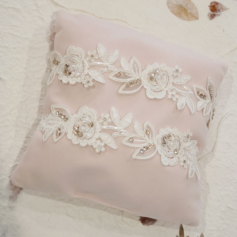 May include: A blush pink pillow with two white lace appliques featuring floral designs and sparkling embellishments.
