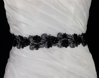 May include: A white dress with a black beaded and lace sash. The sash features a floral design with black beads.