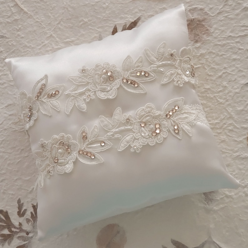 May include: A white satin pillow with two rows of delicate white lace trim adorned with small, sparkling rhinestones.