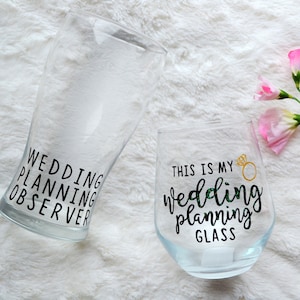May include: Two clear glass cups with black lettering. One cup says "Wedding Planning Observer" and the other says "This is my Wedding Planning Glass" with a gold ring icon.