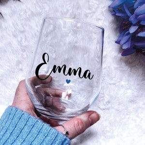 May include: A clear glass wine glass with the name "Emma" written in black cursive lettering. A small blue heart is above the name.