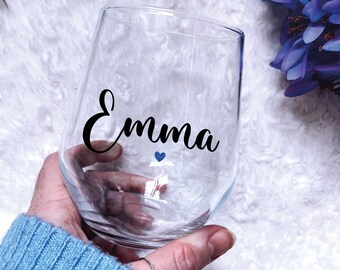 May include: A clear glass wine glass with the name "Emma" written in black cursive lettering. A small blue heart is above the name.