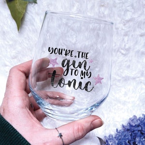 May include: A clear glass wine glass with the text "You're the gin to my tonic" in black lettering with pink glitter stars.
