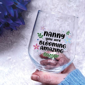 May include: A clear stemless wine glass with the words "Nanny you are blooming amazing" in black script and colorful floral accents. The glass is held in a hand, with a blue sweater sleeve visible.